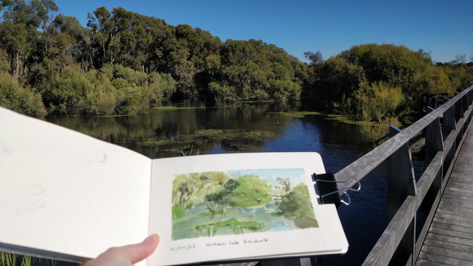 A hand holds out a watercolour landscape painting in a sketchbook with the same view behind of a wetland, with a boardwalk.